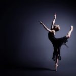 Young ballerina in a black suit is dancing in dark
