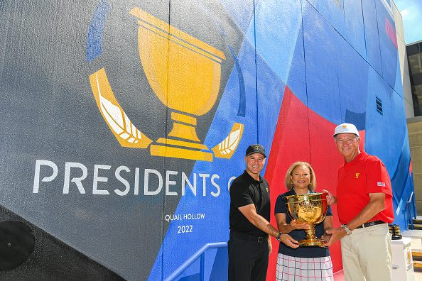 Three people stand holding the President's Cup trophy in front of a mural dedicated to the tournament. 