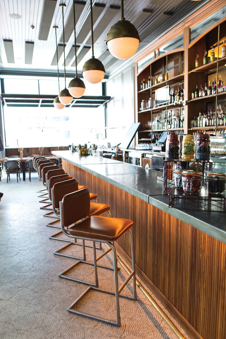 A warmly lit bar with a row of brown leather chairs in front of a gray bar at The Katharine at Kimpton Cardinal Hotel in Winston Salem, NC.