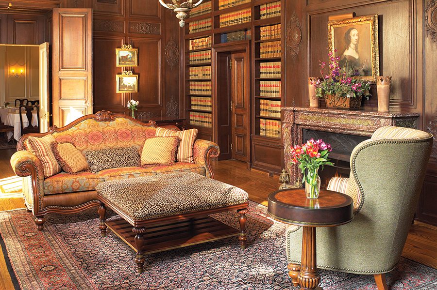 A Victorian decorated room featuring an ornate, oriental couch with various decorative pillows, a animal print patterned ottoman that faces a green upholstered chair that sits next to a wodden side table with flowers , on top of an oriental rug that faces a fireplace with a painted portrait hanging abive it next to a giant floor to ceiling bookshelf at the Graylyn Estate in Winston-Salem, NC.