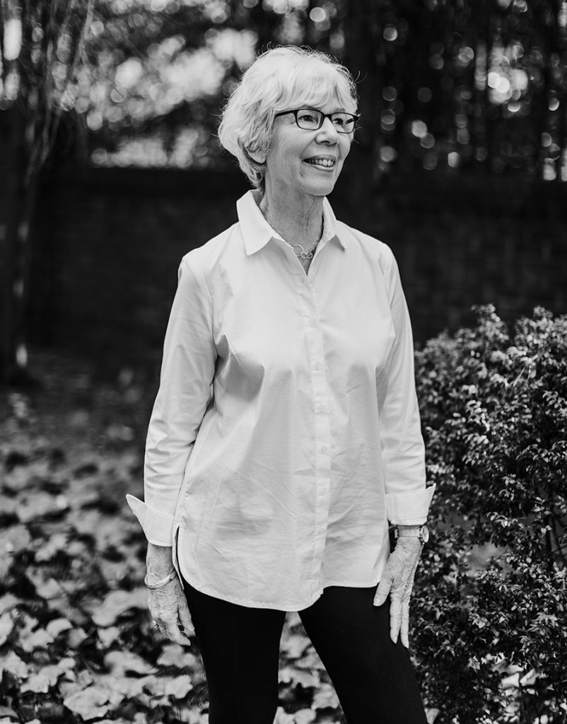 Woman wearing glasses standing in a nature scene wearing a long white button down and black pants with a black and white filter
