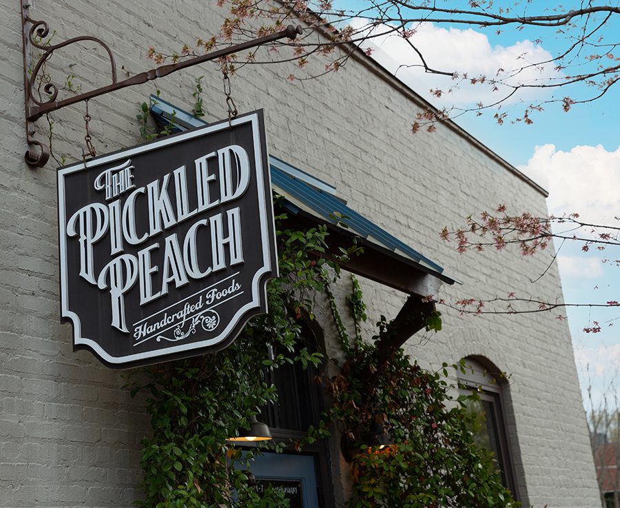 A white painted burck building with cascading ivy and trees surrounding it with a sign that says "The Pickled Peach" in Davidson, NC