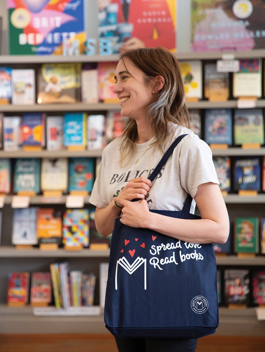 A woman stands in front of a bookshelf holding a tote bag at Main Street Books in Davidson, NC.