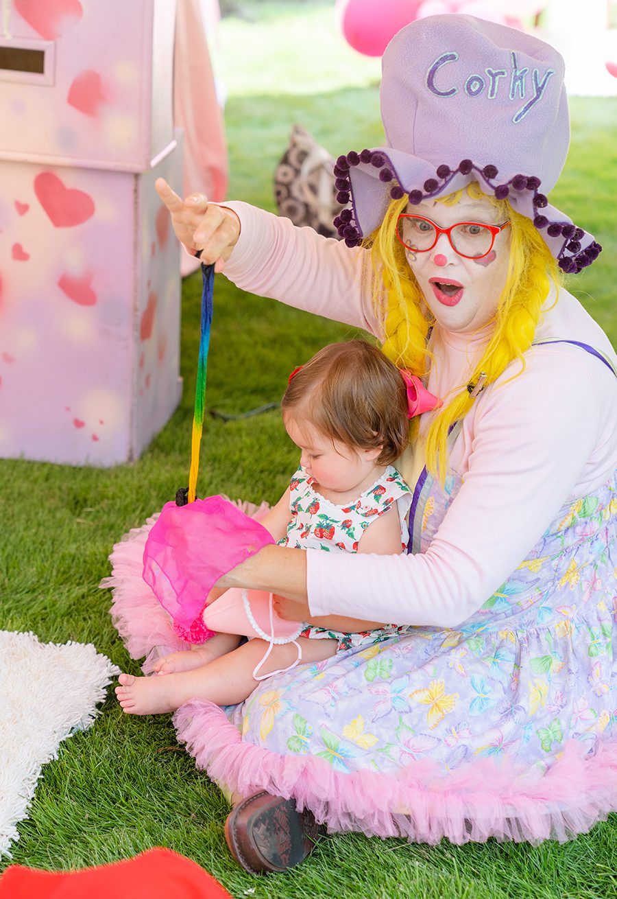 A birthday clown dressed in pastels holds a baby outside while they do a magic trick