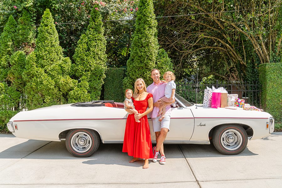 A family of four stands in front of a white vintage Chevrolet convertible wearing reds, pinks, and whites.