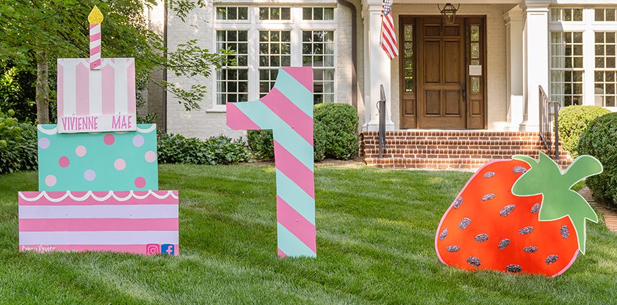 A front yard that showcases a giant pink, teal, and purple painted birthday cake, a pink and teal painted number 1, and a painted strawberry.