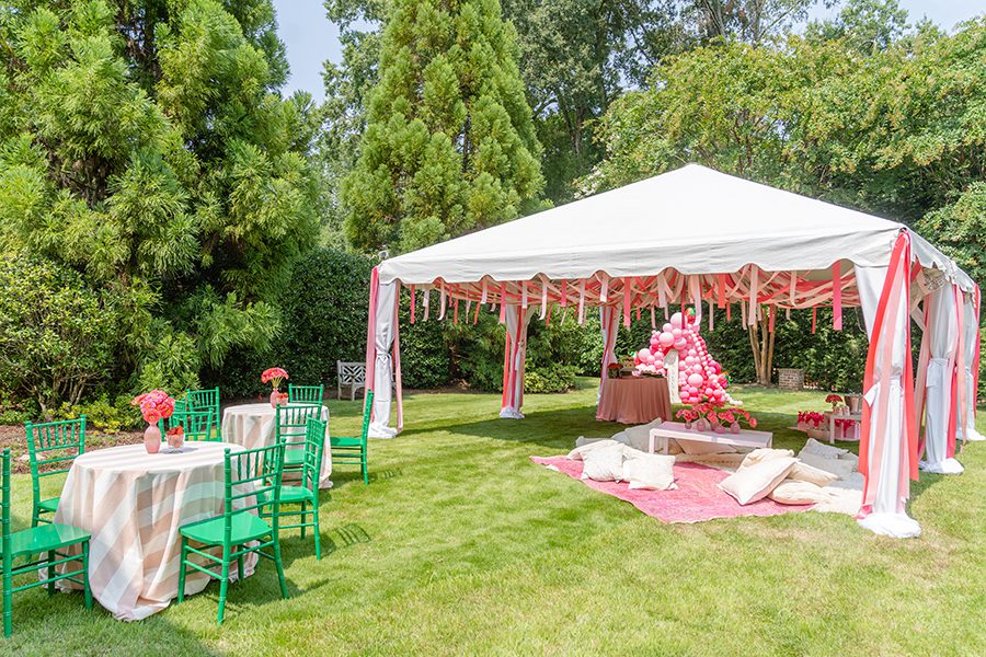 A white tent stands outside decorated with pink streamers and balloons, covering assorted tables and pillows. 