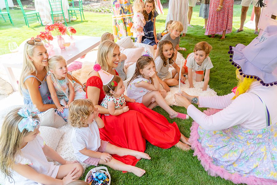 A group of moms with their children sit outside in the grass watching a performance from a birthday clown. 