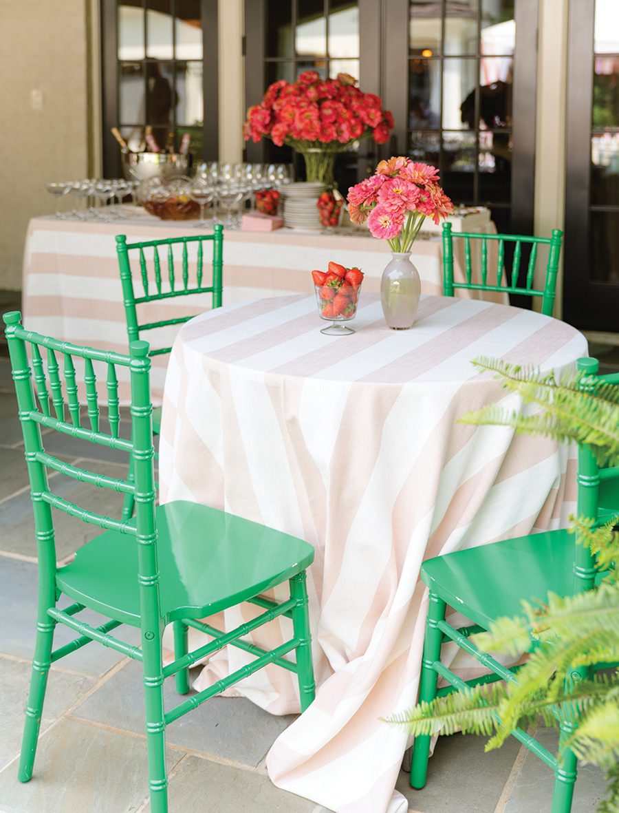 A round table covered with a pink and white striped tablecloth with a vase of pink flowers and a vase of strawberries on top with four seafoam green chairs circling it. 