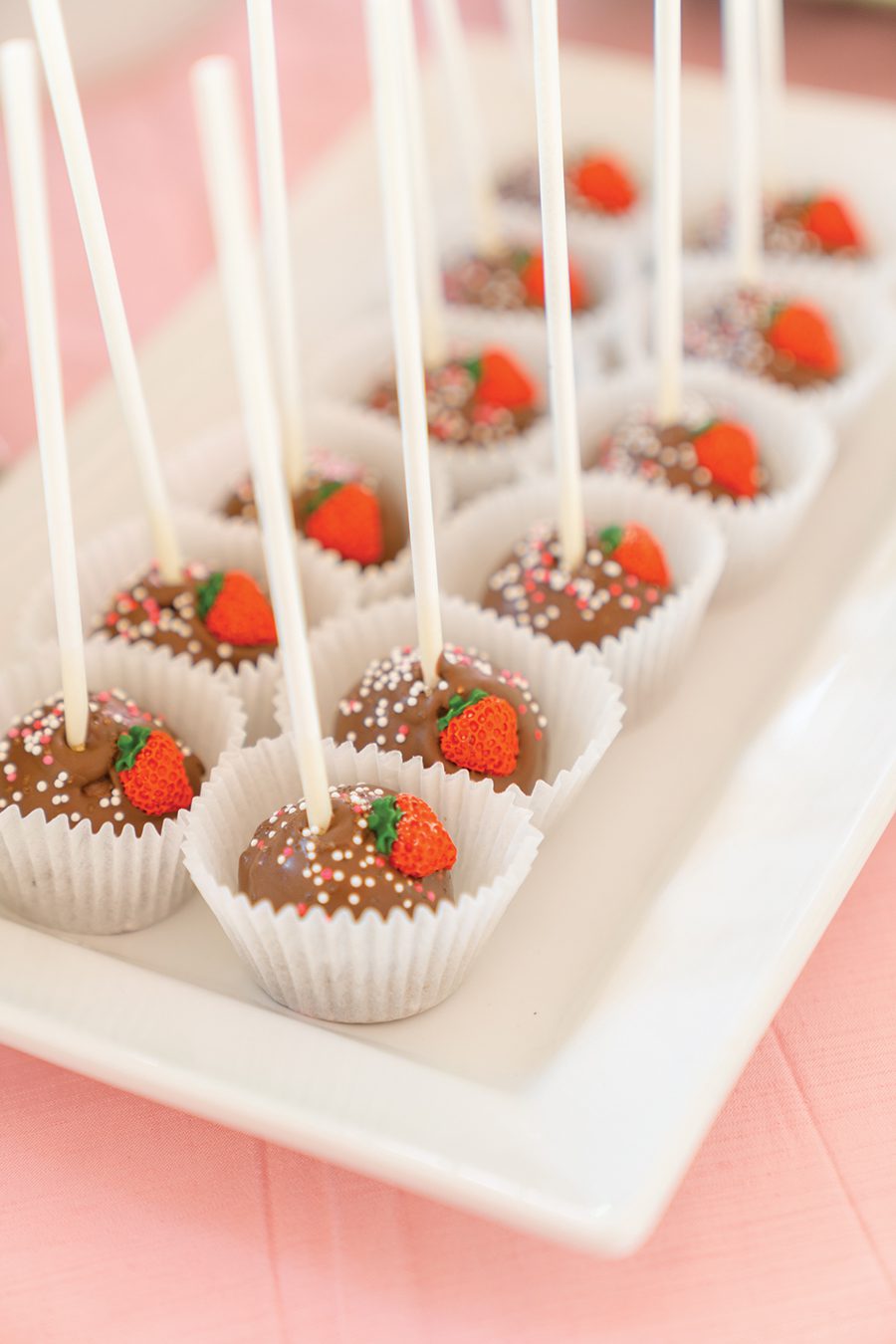 Chocolate covered cake pops with multicolored sprinkles and mini strawberry candies stuck to them. 
