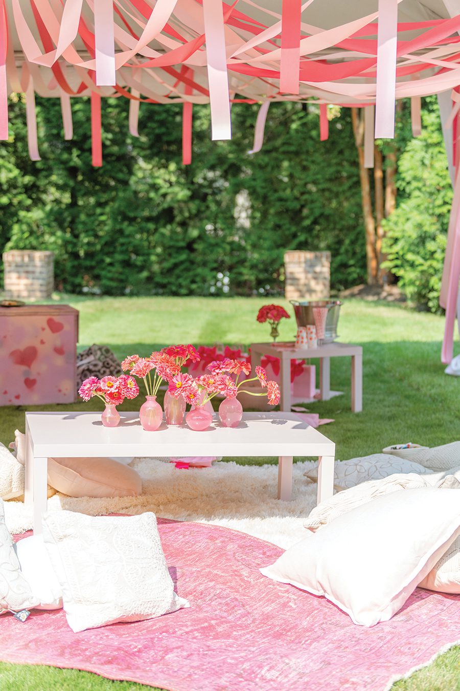 A white table with assorted vases of pink flowers, outside under pink streamers, on top of furry white blankets and white pillows
