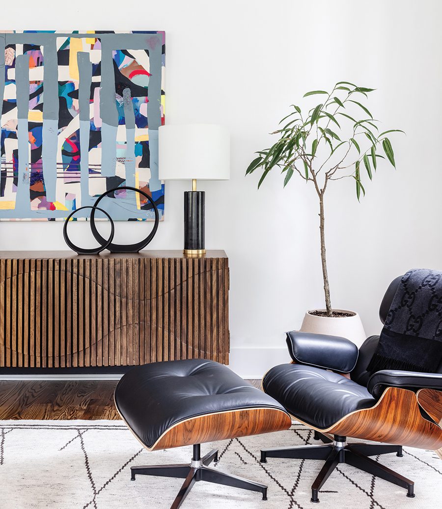 Sunlit room with a colorful abstract painting on the wall, a wooden credenza with black decor and a black lamp, a Bonzai tree in the corner, a black leather chair with a matching ottoman, a white patterned carpet, and hardwood floors. 