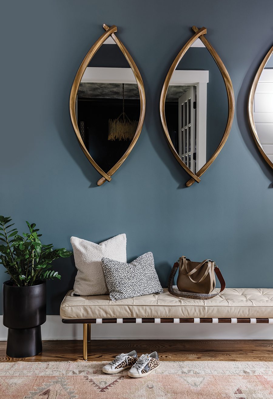 Dark blue green painted walls with three interestingly shaped mirrors, a bench with decorative pillows, and a plant in a small black container.