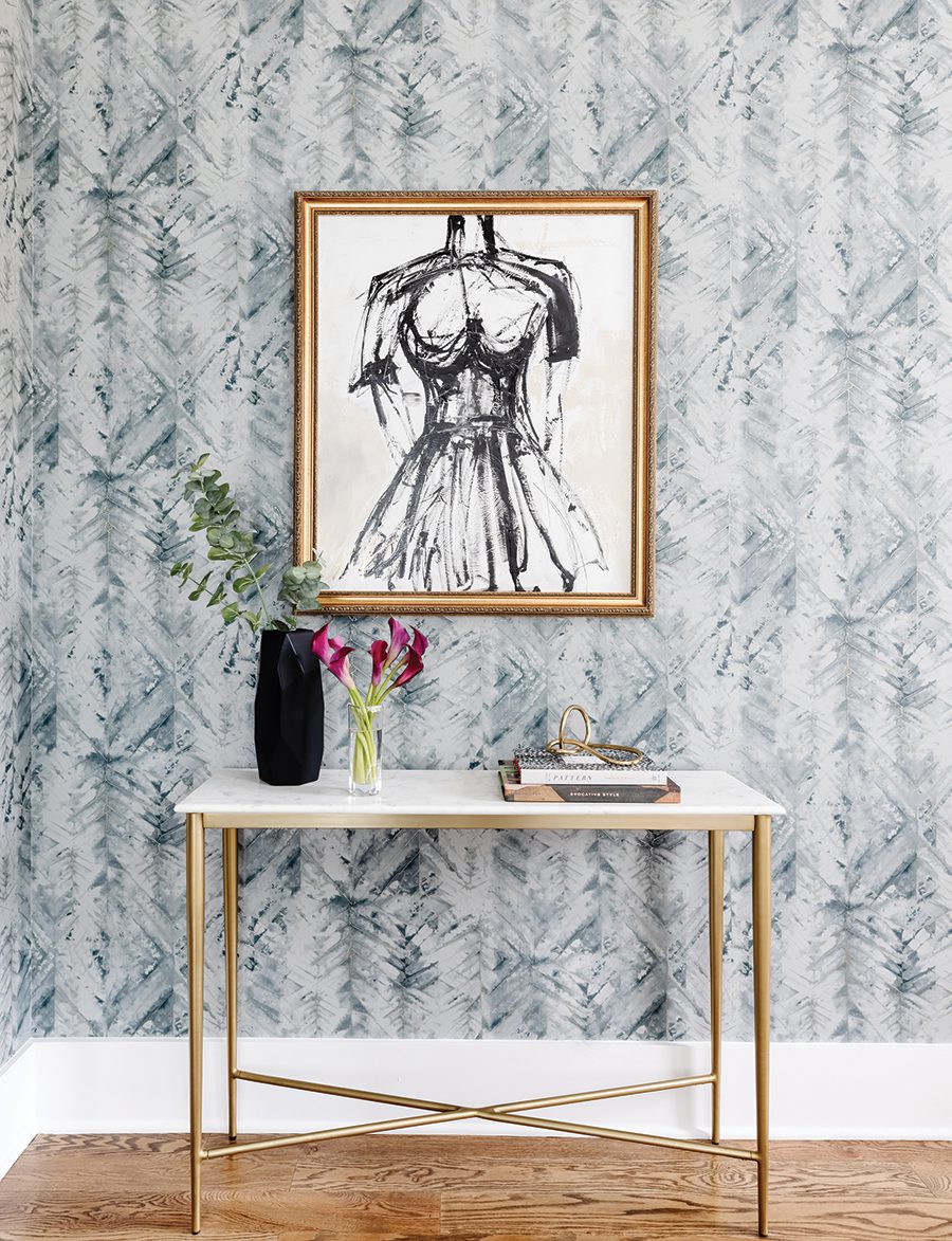 Wallpapered wall with a gold framed art piece of a human form, a white marble top table with assorted coffee table books, plants, and gold decor. 