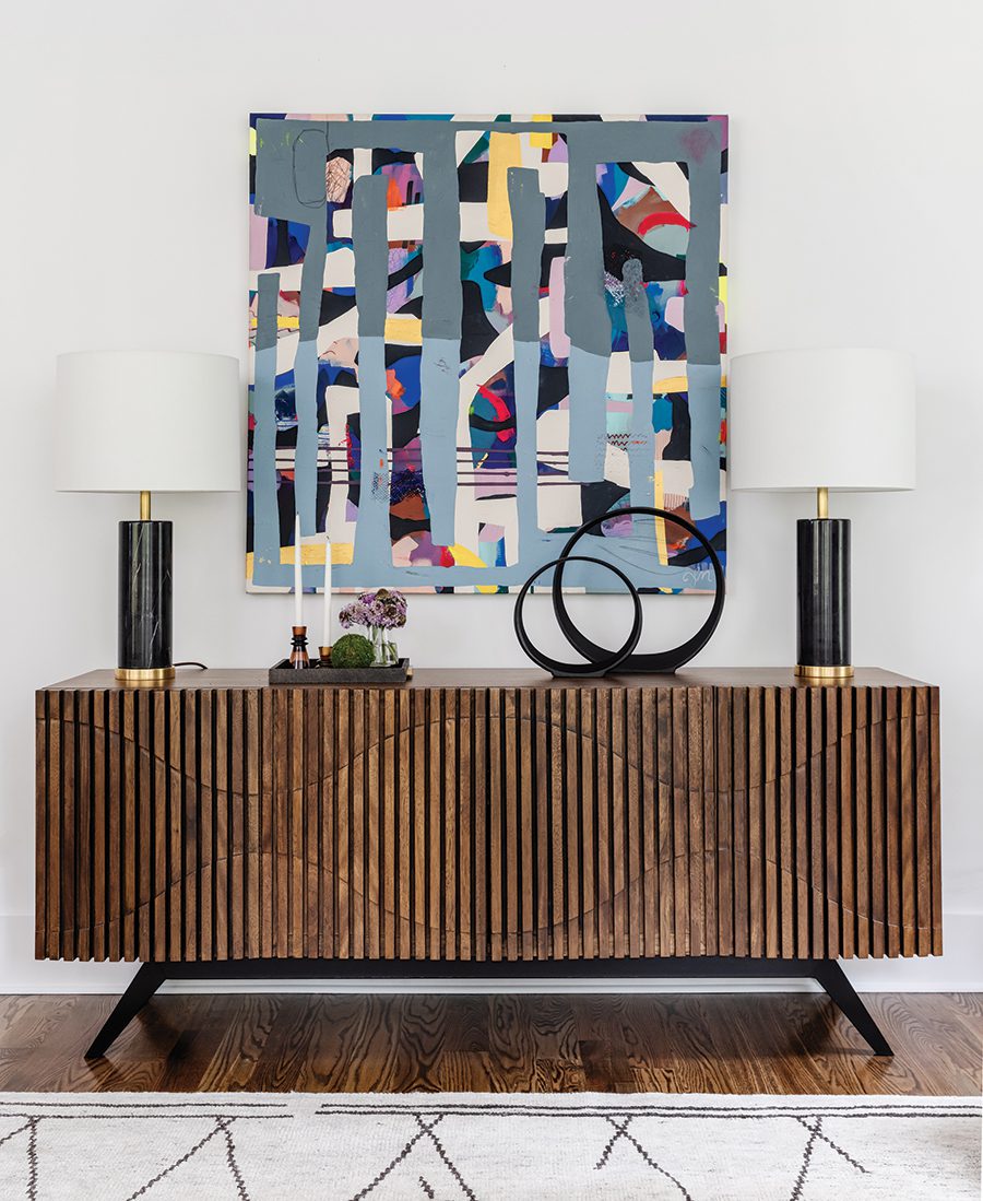 Multi-colored abstract painting hanging on a white wall on top of a wooden credenza with two black lamps and assorted decor.
