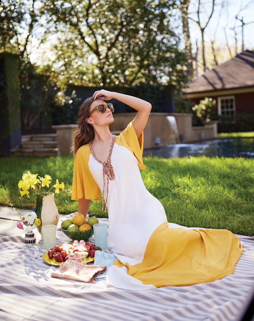 A woman sits outside on a striped picnic blanket wearing a yellow and white dress with sunglasses as she stares into the sky, ignoring the assorted food on the blanket.