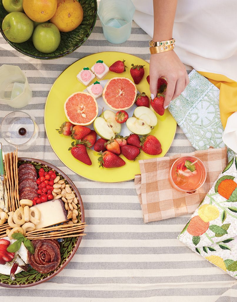 A hand reaches for a strwaberry that is situated on a bright yellow plate amongst other assorted fruits on top of a striped blanket that also holds other items of food as well as drinks and napkins
