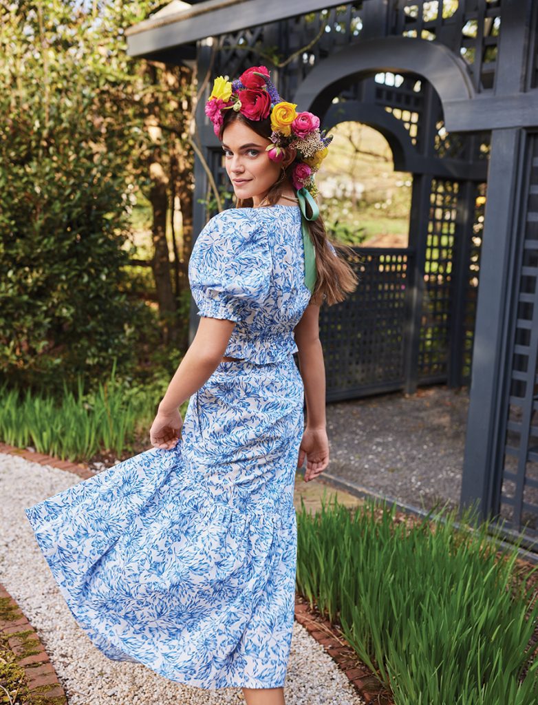 A woman wears a long, blue patterened dress with an intricate floral headpiece while walking towards a gazebo outside along a gravel path