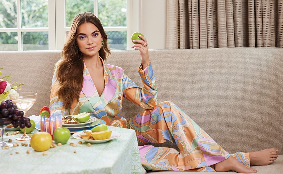 A woman sits on a couch inside wearing a multicolored pastel blouse with matching pants while she holds an apple in front of a table full of food