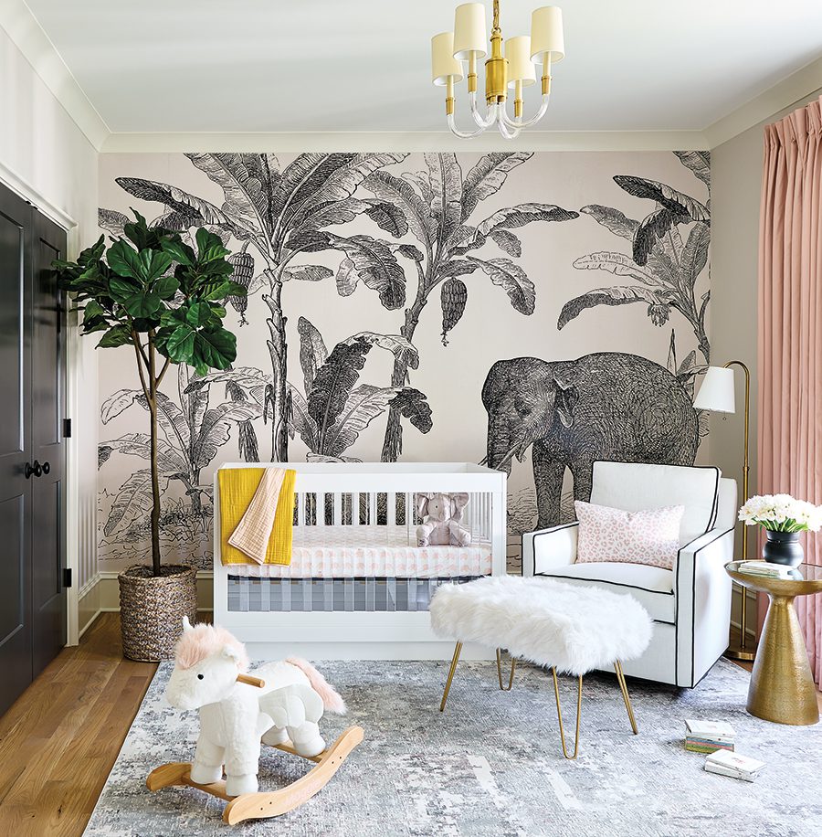 A wallpapered mural that features an elephant with palm trees on a wall facing a white crib with a yellow blanket draped over the side on top of a gray patterned carpet with a white furry ottoman and black-trimmed white chair in front of an overhead lamp next to a gold side table and blush pink curtains.