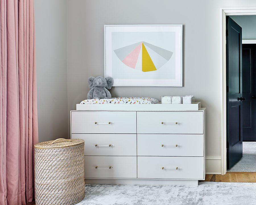 Gray painted wall with a pink, white and gold geometric art piece hanging on to of a white changing station with a stuffed gray elephant on top.