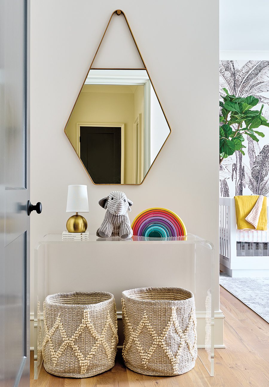 A geometric gold-trimmed mirror hanging on white walls on top of a clear table with a gold lamp, stuffed elephant, and rainbow decor piece with two gray laundry hampers underneath.