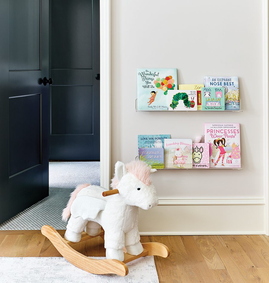 A stuffed rocking unicorn on light hardwood floors in front of a hanging bookshelf with children's books displayed.