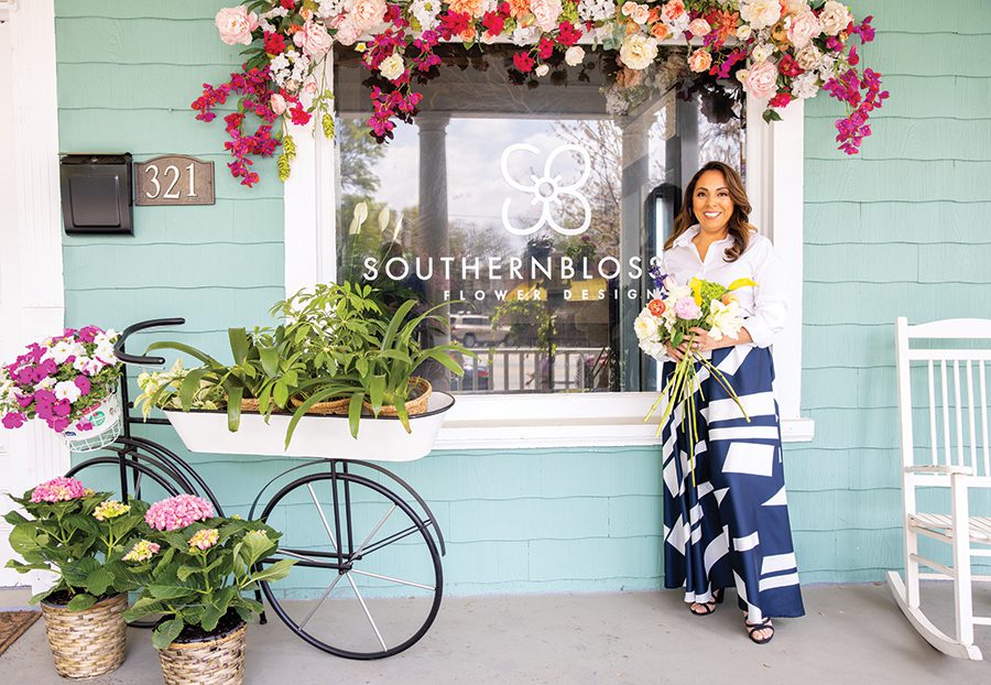 A woman stands in front of a the flower shop Southern Blossom that is painted a light blue color and decorated with multiple different flowers of different colors.