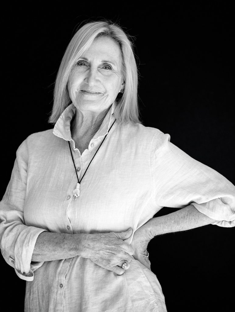 A woman with short white hair stands in front of a black background wearing a white button-down