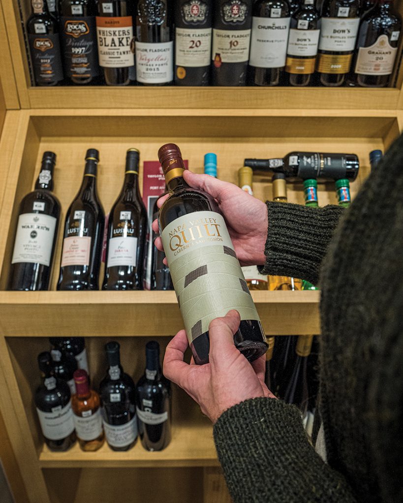 A person holds a bottle of Napa Valley Quilt cabernet in front of a collection of assorted wines