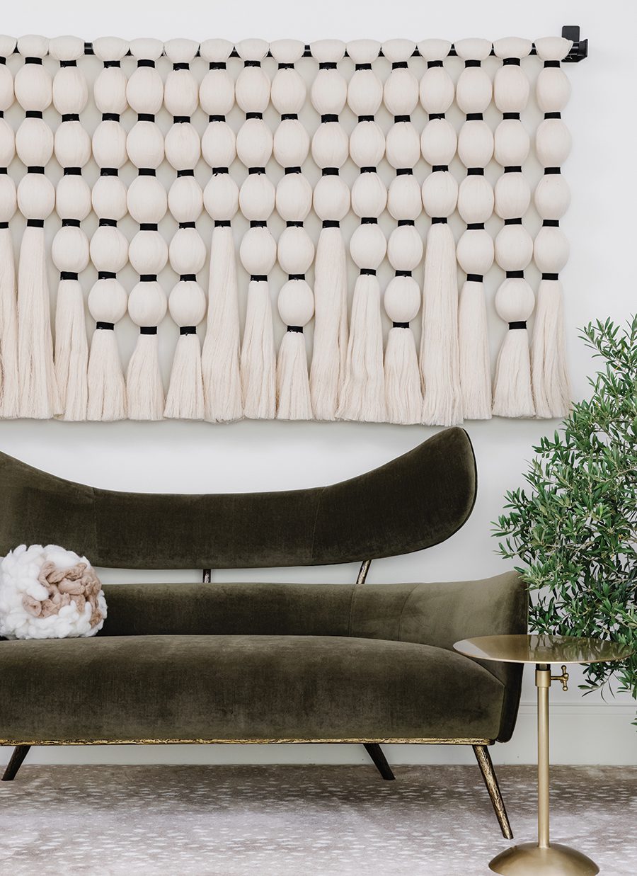An artisan-made white fiber art piece hangs behind a green velvet sofa with a gold side table