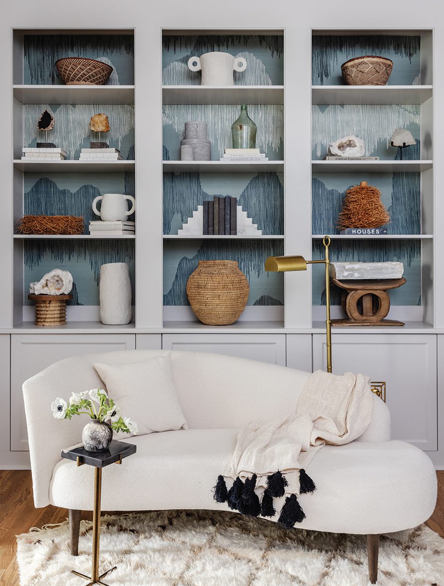 A wallpapered bookshelf displays an array of decor pieces with a white side sofa and gold lamp