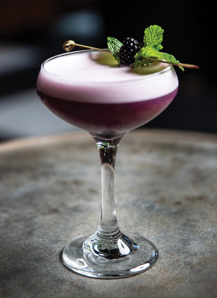 A lavender colored cocktail sits in a cocktail glass with a blackberry garnish on top