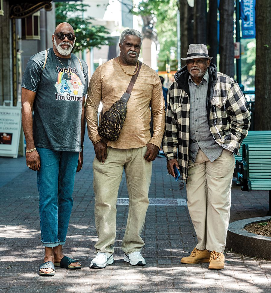 Three men stand on a stone pathway, the one of the far left wears a Care Bears graphic t-shirt jeans and slides. The man in the middle wears a gold chain necklace, a muted yellow long sleeve top, khakis, white Nike sneakers and a Louis Vuitton crossbody fanny pack. The man on the far right wears a gray fedora, gray button down, cream and black plaid coat with khakis and light brown sneakers.