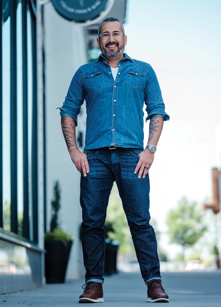 A man stands on a hill wearing a denim button down, black belt, and blue denim jeans with brown sneakers.