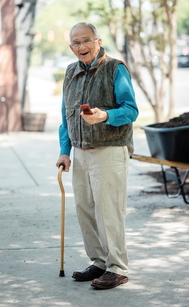 An elderly man stands on the sidewalk holding a cane and his flip phone wearing a blue button down shirt, came zip up vest, khakis, and brown shoes.
