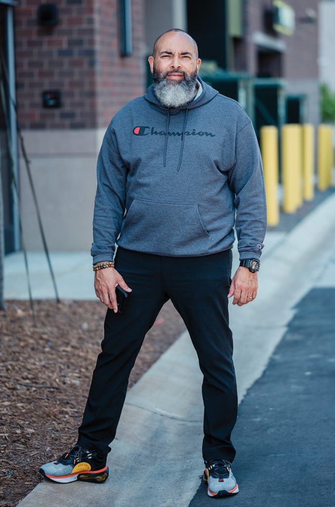 A man stands on a curb wearing a gray Champion sweatshirt, black pants, colorful sneakers, and a wristwatch.