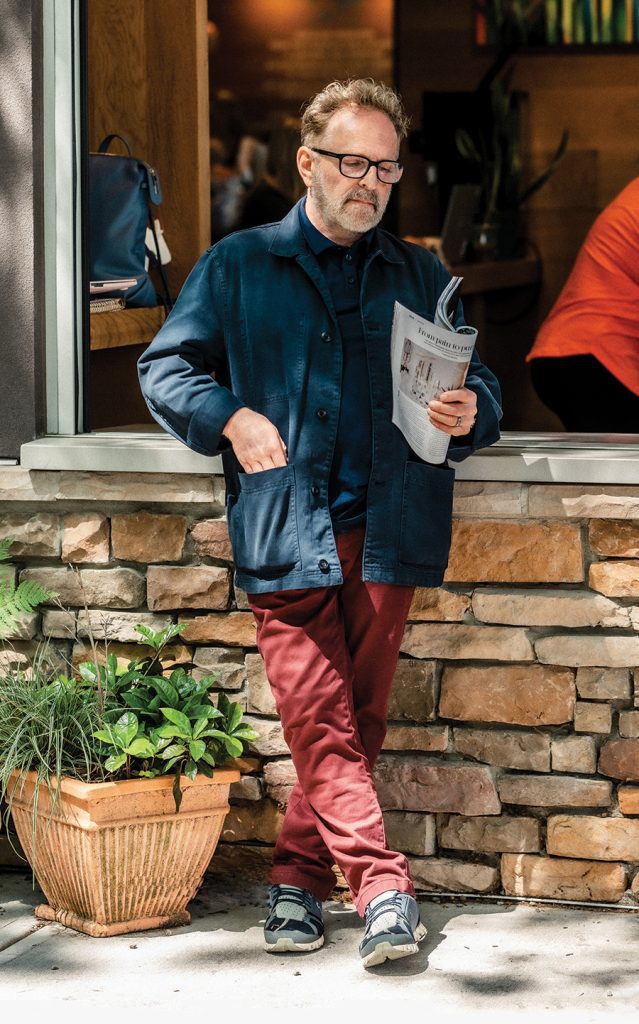 A man leans up against a stone wall holding a magazine wearing a dark colored polo, a dark colored jacket, red pants, and sneakers.
