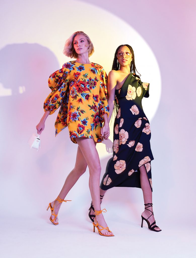 Two women stand in front of a purple background. One wears a yellow floral outfit while holding a small white purse and the other wears a black floral dress.