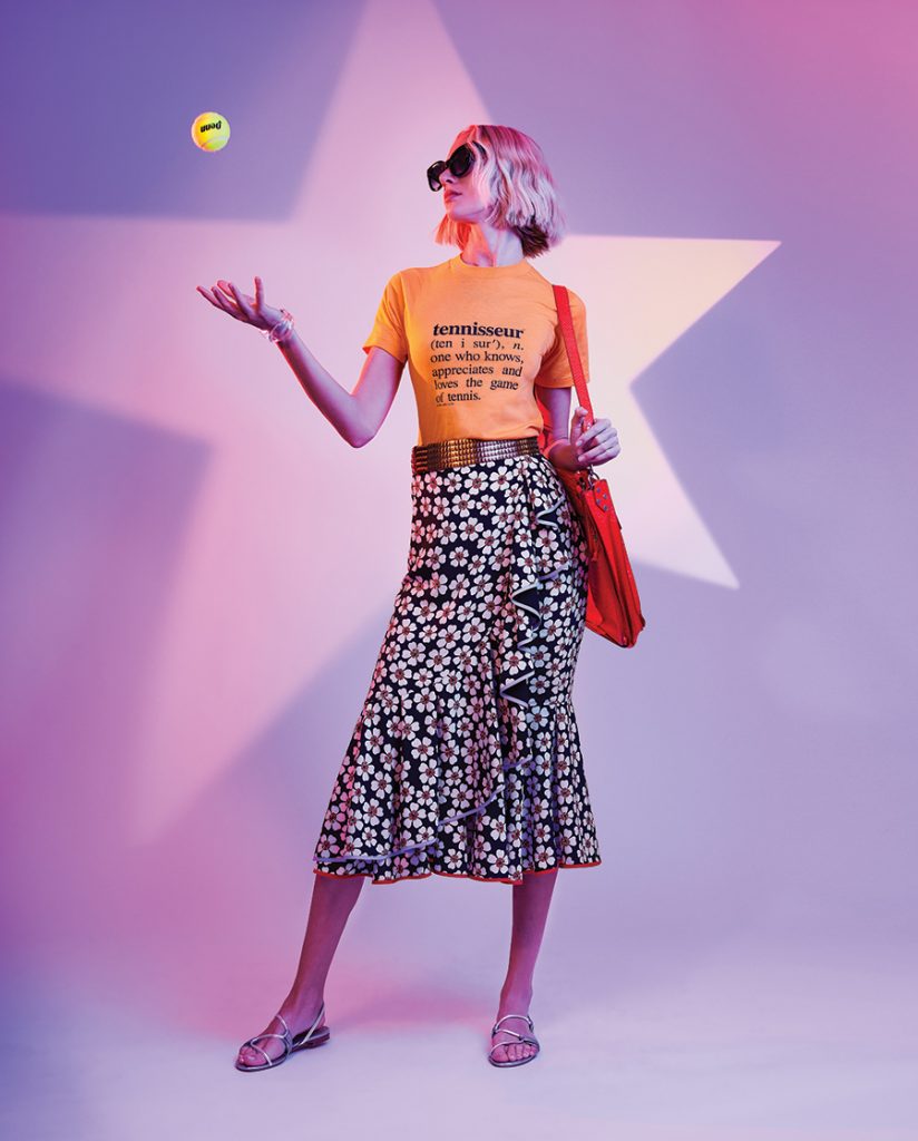A woman stands in front of a purple background with a huge star while wearing a yellow T-shirt, black floral skirt, with a red purse, throwing a tennis ball in the air.