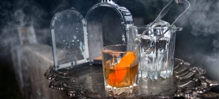 Hallows Cocktail on a decorative silver plate with lantern and crystal glasses. Spooky vapors in the background.