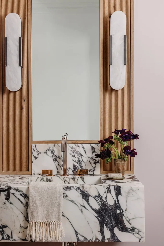 Custom reeded wood accents the vanity in the powder room. A Kohler ombre faucet is nestled into the marble, fabricated by Art of Stone
