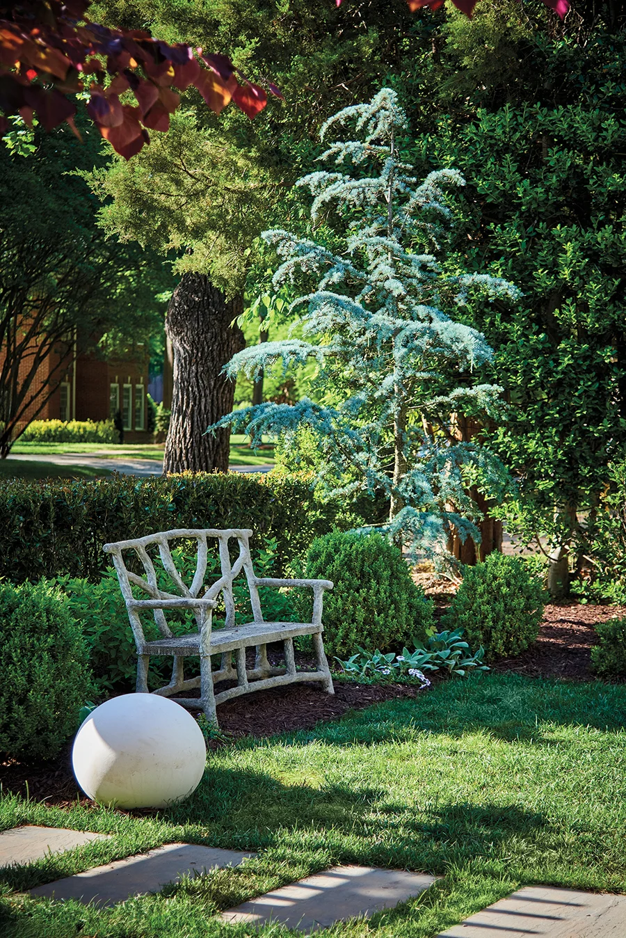 A faux-bois garden bench is nestled between boxwoods and beneath a Blue Atlas cedar tree in the corner of the front garden.
