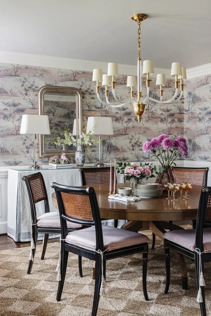 “Sagimai,” a printed grasscloth wallpaper by Scalamandre, creates a soothing backdrop in the dining room. The chairs and table are by Jonathan Charles. A side table with a custom table skirt provides needed storage without taking up too much space. The chandelier is “Vivian” by Visual Comfort, and the mirror is from Granville.